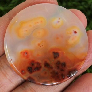 Peut inclure: Une pierre précieuse ronde et polie, présentant un mélange de couleurs. La pierre a une base blanc crème avec des motifs orange et rouge. Les motifs comprennent des taches et des tourbillons, créant un design unique. La pierre est tenue dans une main.
