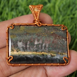 May include: A rectangular pendant with a natural stone center, wrapped in copper wire. The stone features a mix of green, brown, and gold hues. The wirework forms a frame and a bail for a necklace. The pendant is held in a hand.