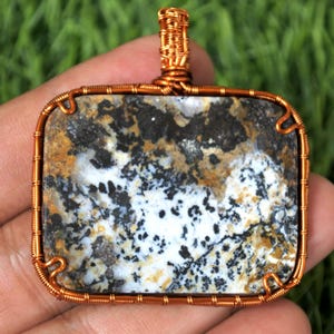 Puede incluir: Colgante rectangular con un centro de piedra natural, con una mezcla de patrones negros, blancos y tostados. La piedra está encerrada en un marco y una fianza de alambre de cobre, creando una estética rústica y artesanal. El colgante es probablemente para joyería.