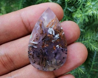 May include: A teardrop-shaped gemstone with a mix of colors. The stone features a translucent lavender base with brown, white, and black inclusions. The polished surface of the stone is smooth and reflective, showcasing the natural patterns.