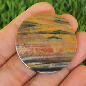 Puede incluir: Una piedra redonda y plana, multicolor, con un patrón en capas de negro, blanco, amarillo, naranja y rojo. La piedra tiene una superficie lisa y pulida.