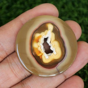 May include: A round, polished agate stone with a natural hole in the centre. The stone has a brown, yellow, and white colour pattern.