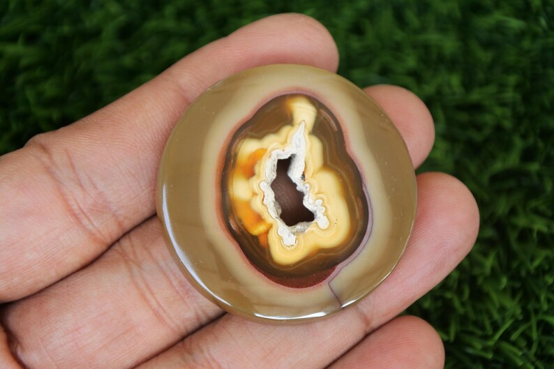 May include: A round, polished agate stone with a natural hole in the centre. The stone has a brown, yellow, and white colour pattern.
