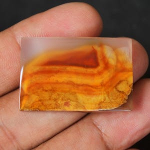 May include: A rectangular, polished agate cabochon with a translucent, white base and a layered pattern of orange, yellow, and brown.