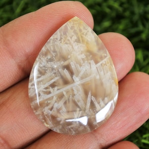 Könnte beinhalten: Ein tropfenförmiger, polierter Edelstein. Der Stein zeigt beige und weiße Muster, die eine natürliche Landschaft darstellen. Der Edelstein wird in einer Hand gehalten, um seine Größe und Form zu zeigen. Geeignet für die Schmuckherstellung.