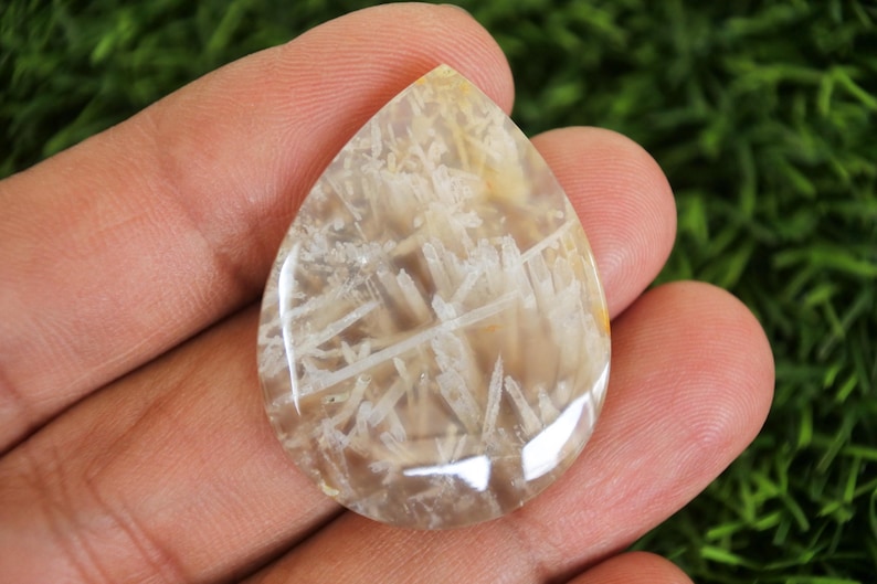 Peut inclure: Une pierre pr&eacute;cieuse polie en forme de goutte d'eau. La pierre pr&eacute;sente des motifs beiges et blancs, &eacute;voquant un paysage naturel. Le bijou est tenu dans une main, mettant en valeur sa taille et sa forme. Id&eacute;al pour la cr&eacute;ation de bijoux.