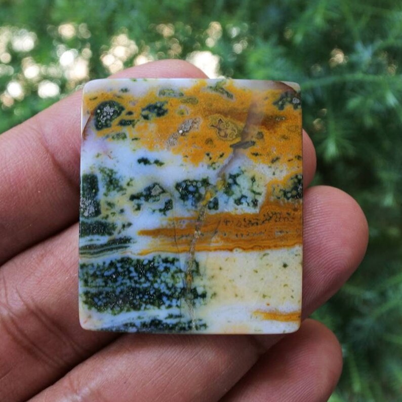 May include: A rectangular gemstone with a polished surface. The stone features a blend of colours, including yellow, green, white, and brown, creating a unique pattern. The gemstone is held in a hand, with a blurred green background.