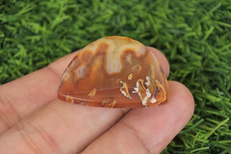 May include: A brown and white agate cabochon with a unique pattern. The cabochon is smooth and polished, with a slightly rounded shape.