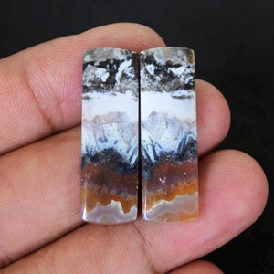 Könnte beinhalten: Zwei rechteckige Edelsteine mit geschichtetem Aussehen. Der obere Abschnitt ist eine Mischung aus Schwarz und Weiß, gefolgt von Weiß, dann Blau und Grau und schließlich einer braunen und orangefarbenen Basis. Die Steine werden in einer Hand gehalten.