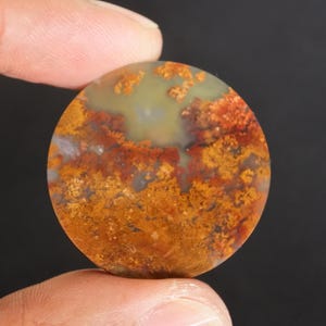 Puede incluir: Una gema redonda y pulida con un patr&oacute;n &uacute;nico de inclusiones naranjas, rojas y amarillas. La piedra tiene un color base verde claro y blanco. La gema se sostiene entre dos dedos sobre un fondo oscuro.