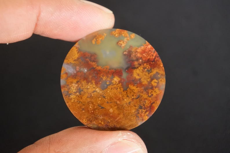 Puede incluir: Una gema redonda y pulida con un patr&oacute;n &uacute;nico de inclusiones naranjas, rojas y amarillas. La piedra tiene un color base verde claro y blanco. La gema se sostiene entre dos dedos sobre un fondo oscuro.