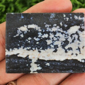 May include: A square, polished stone with a striking pattern. The stone features a dark, almost black base color, contrasted by white and light beige cloud-like formations. The overall effect is a unique, natural design, perfect for jewelry or decorative use.