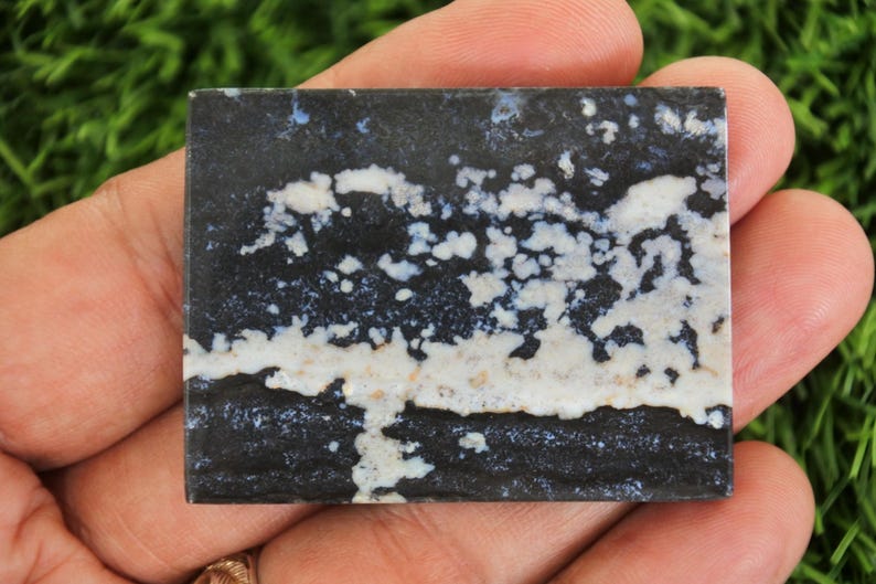 May include: A square, polished stone with a striking pattern. The stone features a dark, almost black base color, contrasted by white and light beige cloud-like formations. The overall effect is a unique, natural design, perfect for jewelry or decorative use.