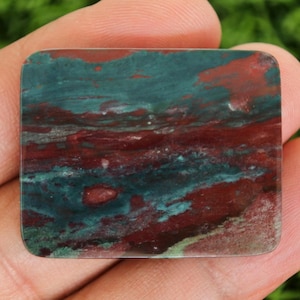 May include: A rectangular gemstone with a polished surface. The stone displays a mix of deep red and teal green colours, creating a marbled effect. The colours are swirled and layered, giving the stone a unique, natural appearance. The gemstone is held in a hand.