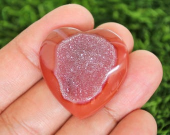 Puede incluir: Una gema pulida en forma de corazón, de color rojo anaranjado. El centro presenta una formación cristalina brillante en tonos rosa y blanco. Este artículo decorativo es único.