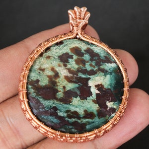 Peut inclure: Un pendentif en fil de cuivre enveloppé avec une pierre ronde verte et brune avec un motif tourbillonnant.