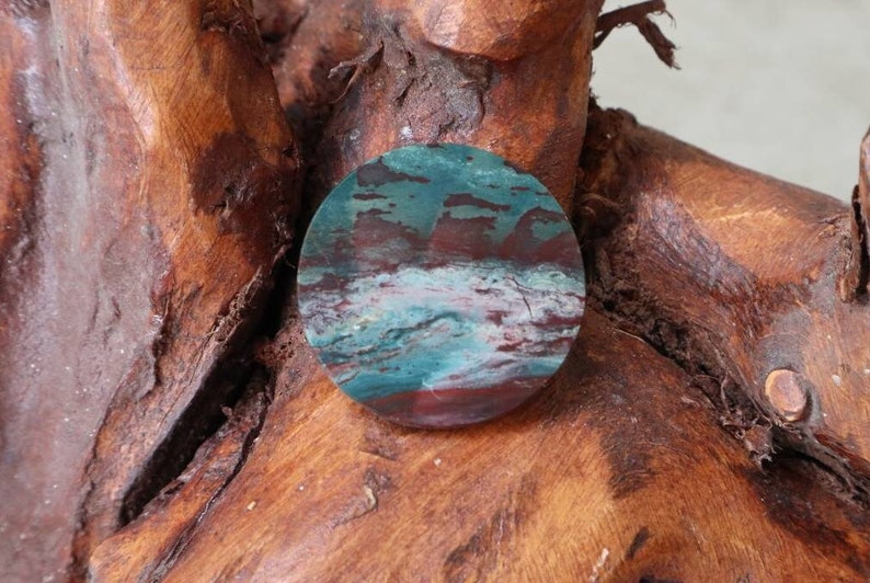 May include: A round, polished stone with a swirling pattern of blue, green, and red colours. The stone is set against a brown wooden background.