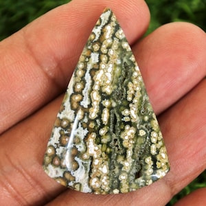 Puede incluir: Una gema triangular con un patrón único de manchas y líneas verdes, blancas y marrones. La piedra pulida se sostiene en una mano, mostrando sus colores y texturas naturales. La forma de la piedra recuerda a un pequeño árbol de Navidad estilizado.