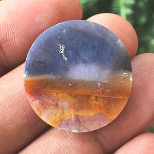 May include: A circular, polished stone displaying a natural landscape pattern. The upper section is a deep blue, fading into white, then a band of orange and red. The stone has a smooth, glossy finish. It is held in a hand.