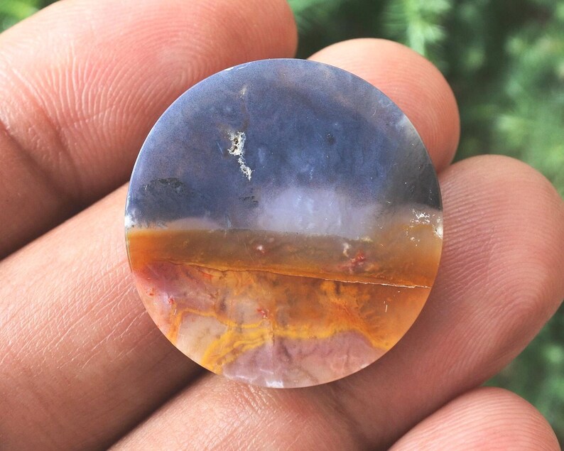 May include: A circular, polished stone displaying a natural landscape pattern. The upper section is a deep blue, fading into white, then a band of orange and red. The stone has a smooth, glossy finish. It is held in a hand.