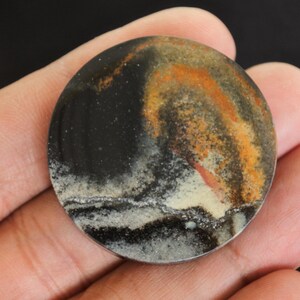 K&ouml;nnte beinhalten: Ein runder, polierter Stein mit einem einzigartigen Muster aus schwarzen, orangefarbenen und wei&szlig;en Wirbeln. Der Stein wird in einer Hand gehalten, wodurch seine nat&uuml;rlichen Farben und Texturen hervorgehoben werden. Das Design &auml;hnelt einer Miniaturlandschaft.