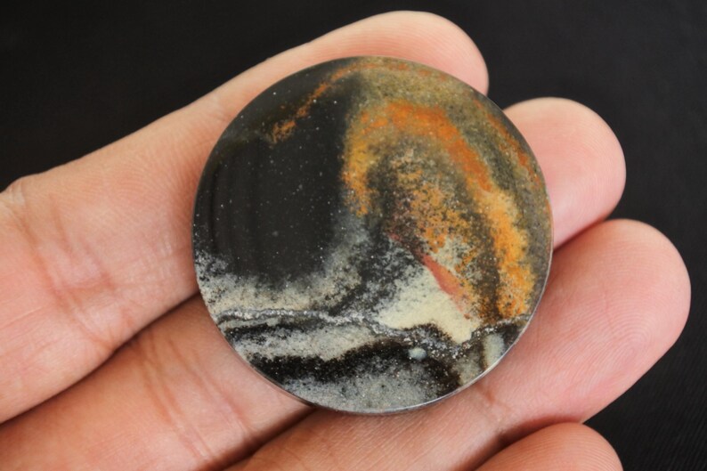 K&ouml;nnte beinhalten: Ein runder, polierter Stein mit einem einzigartigen Muster aus schwarzen, orangefarbenen und wei&szlig;en Wirbeln. Der Stein wird in einer Hand gehalten, wodurch seine nat&uuml;rlichen Farben und Texturen hervorgehoben werden. Das Design &auml;hnelt einer Miniaturlandschaft.