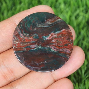 Może przedstawiać: Okrągły, polerowany kamień z wirującym wzorem w odcieniach głębokiej czerwieni, ciemnej zieleni i brązu. Powierzchnia kamienia ma błyszczące wykończenie, a kolory łączą się w naturalny, organiczny wzór. Ten dekoracyjny kamień jest idealny do tworzenia biżuterii lub ekspozycji.