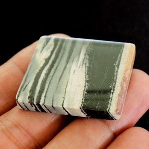 May include: A square stone with a layered pattern of green, white, and black. The stone has a polished surface and a natural appearance. The edges show a mix of colours, including tan and reddish-brown.