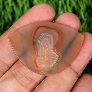 May include: A translucent, triangular-shaped agate stone with a layered pattern of orange, white, and red hues. The stone is held in a hand, showcasing its natural beauty and unique design. The agate is a decorative gemstone.