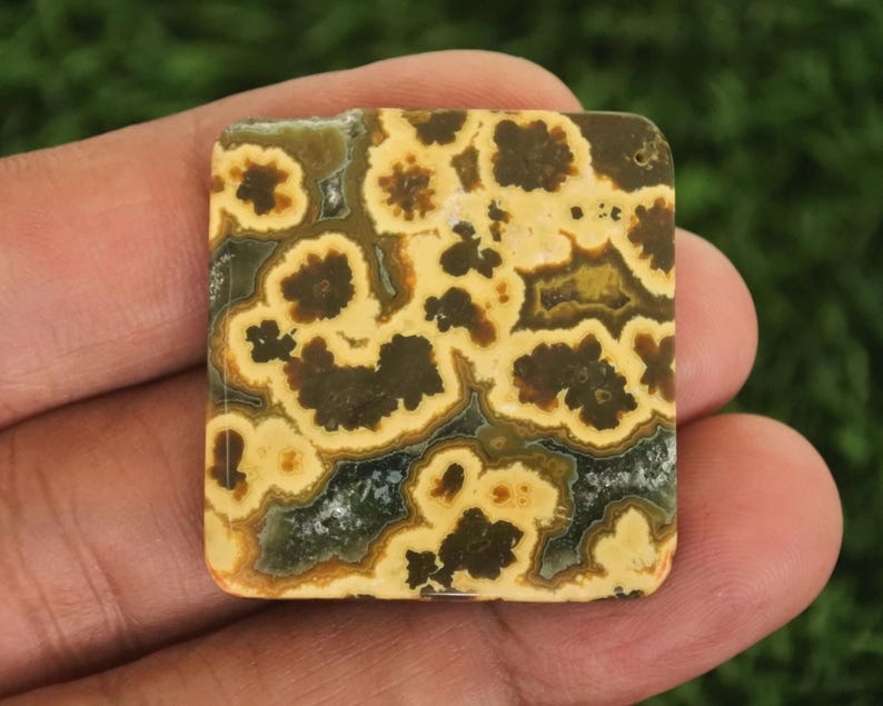 Peut inclure: Une pierre pr&eacute;cieuse carr&eacute;e avec un motif unique de couleurs marron, jaune et cr&egrave;me. La surface polie pr&eacute;sente un aspect naturel et organique. Le design complexe ressemble &agrave; une mosa&iuml;que ou &agrave; un paysage abstrait, d'environ 2,5 cm.