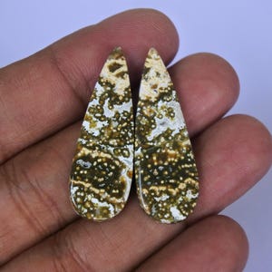 May include: Two teardrop-shaped gemstones with a unique pattern. The stones feature a mix of olive green, cream, and brown colors, with a speckled appearance. The gemstones are held in a hand.