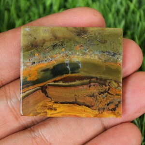 May include: Square-shaped gemstone with a layered pattern of earthy tones. The stone features bands of brown, yellow, and green, creating a natural, textured appearance. The gemstone is held in a hand, showcasing its polished surface.
