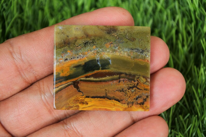 May include: Square-shaped gemstone with a layered pattern of earthy tones. The stone features bands of brown, yellow, and green, creating a natural, textured appearance. The gemstone is held in a hand, showcasing its polished surface.