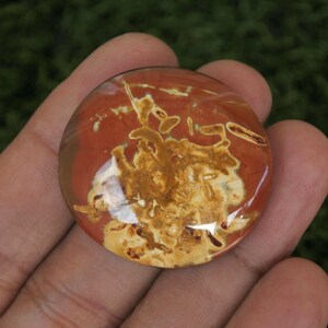 Può includere: Un cabochon rotondo e traslucido con una base rossa e motivi intricati e vorticosi in tonalità di marrone e bianco.