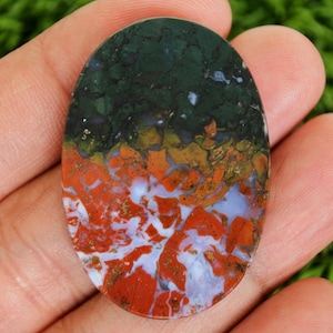 May include: Oval-shaped gemstone with a striking color pattern. The top portion is dark green, transitioning to orange and red with white and blue veins. The polished stone is held in a hand, showcasing its natural beauty.