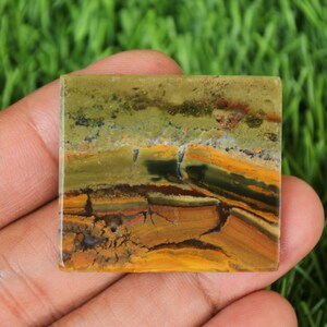 May include: Square-shaped stone with a layered pattern of earthy tones. The stone features bands of olive green, brown, and golden yellow, creating a natural, textured appearance. The stone is held in a hand, showcasing its polished surface.