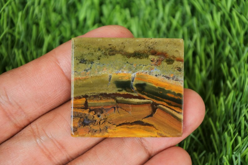 May include: Square-shaped stone with a layered pattern of earthy tones. The stone features bands of olive green, brown, and golden yellow, creating a natural, textured appearance. The stone is held in a hand, showcasing its polished surface.