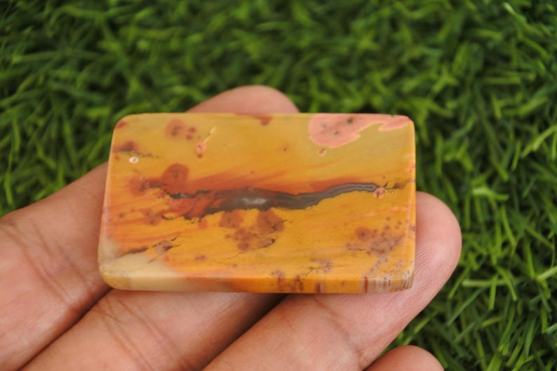 May include: A rectangular polished gemstone cabochon featuring a warm color palette of mustard yellow, burnt orange, and cream. The stone displays natural organic inclusions, including dark brown dendritic patterns and soft pinkish-peach patches.