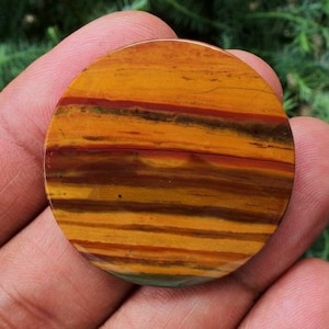 Puede incluir: Una piedra redonda y pulida con un llamativo patrón de bandas horizontales en tonos amarillos, naranjas, rojos y marrones. La superficie lisa de la piedra muestra variaciones naturales de color y textura, lo que sugiere que es una gema o un elemento decorativo. Diámetro aproximado de 5 cm.
