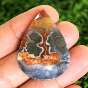 Może przedstawiać: Klejnot w kształcie łzy z unikalnym wzorem kolorów. Kamień charakteryzuje się mieszanką odcieni pomarańczowego, brązowego, szarego i białego, z marmurkowym wyglądem. Wypolerowana powierzchnia odbija światło, podkreślając naturalne wariacje kamienia.