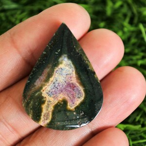 May include: A teardrop-shaped gemstone with a dark green exterior and a central pattern of beige, pink, and purple hues. The polished stone is held in a hand, showcasing its natural colors and unique design.