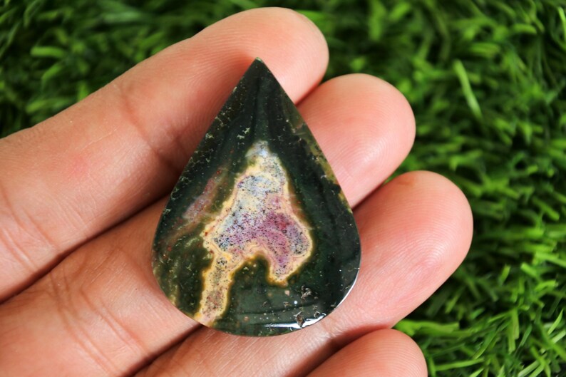 May include: A teardrop-shaped gemstone with a dark green exterior and a central pattern of beige, pink, and purple hues. The polished stone is held in a hand, showcasing its natural colors and unique design.