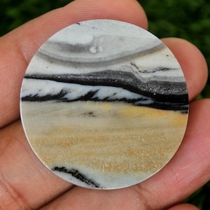 May include: A circular, polished stone displaying a layered pattern of white, grey, black, and tan shades. The stone's surface has a natural, organic look, with bands of colour resembling a landscape. The stone is held in a hand.