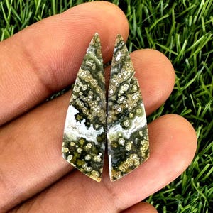 Puede incluir: Dos colgantes de piedras preciosas pulidas con un patrón único. Las piedras presentan una mezcla de colores verde oliva, blanco y marrón, con un aspecto moteado y veteado. Los colgantes tienen forma de lágrima, con la parte posterior plana y la parte frontal pulida.