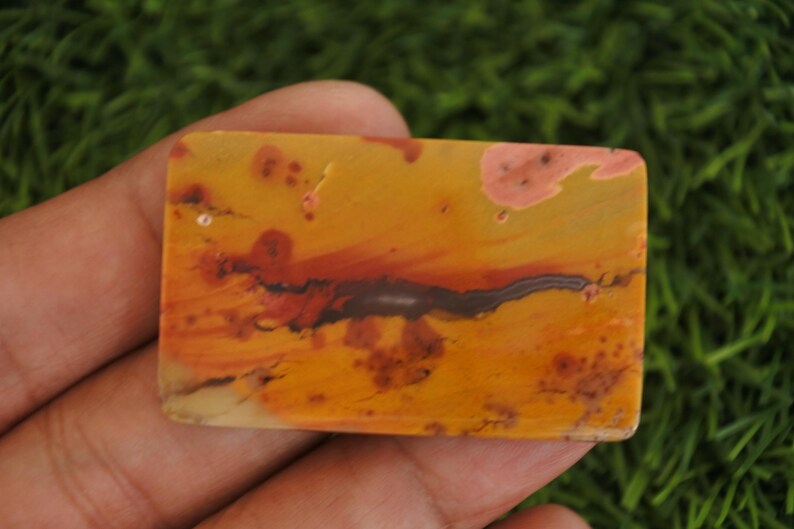 May include: A rectangular polished gemstone cabochon featuring a warm color palette of mustard yellow, burnt orange, and deep red. The stone displays natural organic patterns with dark brown linear inclusions and soft pinkish-tan mottled spots.