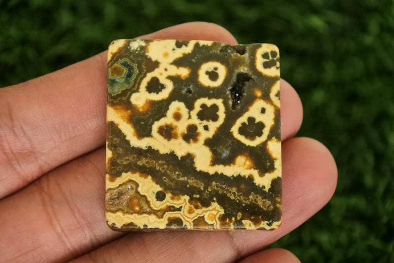 Puede incluir: Gema cuadrada con un patr&oacute;n &uacute;nico de remolinos y manchas de color beige, marr&oacute;n y verde oscuro. La piedra se sostiene en una mano, mostrando sus colores y texturas naturales. Esta piedra pulida podr&iacute;a usarse para joyer&iacute;a u otras manualidades.