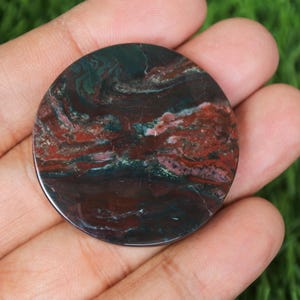 May include: A round, polished stone with a swirling pattern of deep red, dark green, and brown hues. The stone is held in a hand against a backdrop of green grass. The polished surface reflects light, highlighting the natural variations in color and texture.