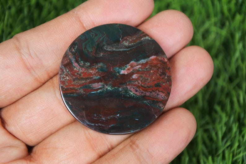 May include: A round, polished stone with a swirling pattern of deep red, dark green, and brown hues. The stone is held in a hand against a backdrop of green grass. The polished surface reflects light, highlighting the natural variations in color and texture.