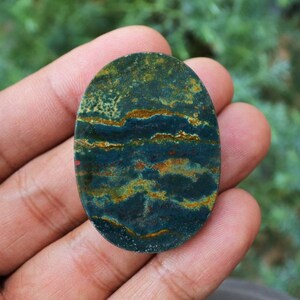 K&ouml;nnte beinhalten: Ein ovaler Edelstein mit einem einzigartigen Muster aus dunkelblaugr&uuml;nen, goldenen und rostfarbenen B&auml;ndern. Die polierte Oberfl&auml;che des Steins zeigt ein nat&uuml;rliches, geschichtetes Aussehen mit Farb- und Texturvariationen.