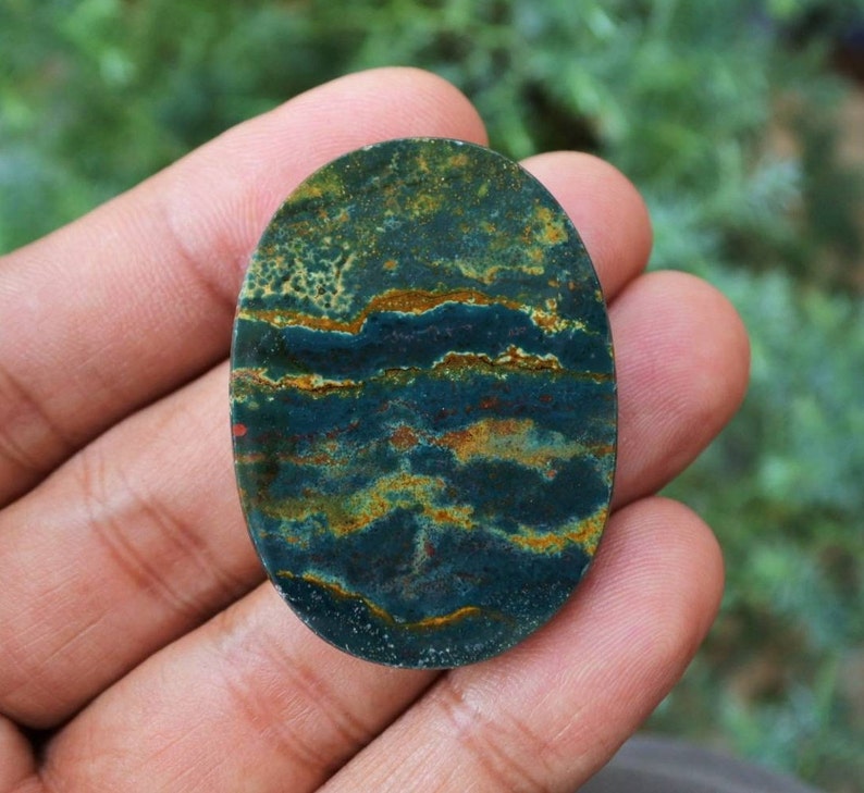 K&ouml;nnte beinhalten: Ein ovaler Edelstein mit einem einzigartigen Muster aus dunkelblaugr&uuml;nen, goldenen und rostfarbenen B&auml;ndern. Die polierte Oberfl&auml;che des Steins zeigt ein nat&uuml;rliches, geschichtetes Aussehen mit Farb- und Texturvariationen.